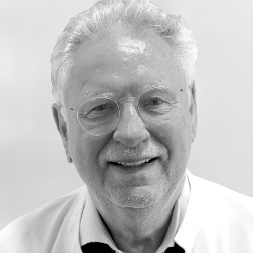 Ein älterer Herr mit grauen Haaren und Brille lächelt freundlich. Er trägt einen weißen Kittel, der auf einen medizinischen oder professionellen Hintergrund hinweist. Der Hintergrund ist unscharf und hell, was den Fokus auf sein Gesicht legt.