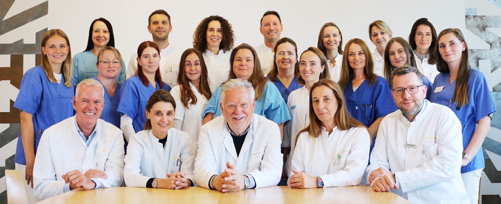 A diverse group of healthcare professionals poses together. They are dressed in white and blue medical attire, with some seated and others standing behind them. The setting appears to be a modern medical facility, conveying a sense of teamwork and professionalism.