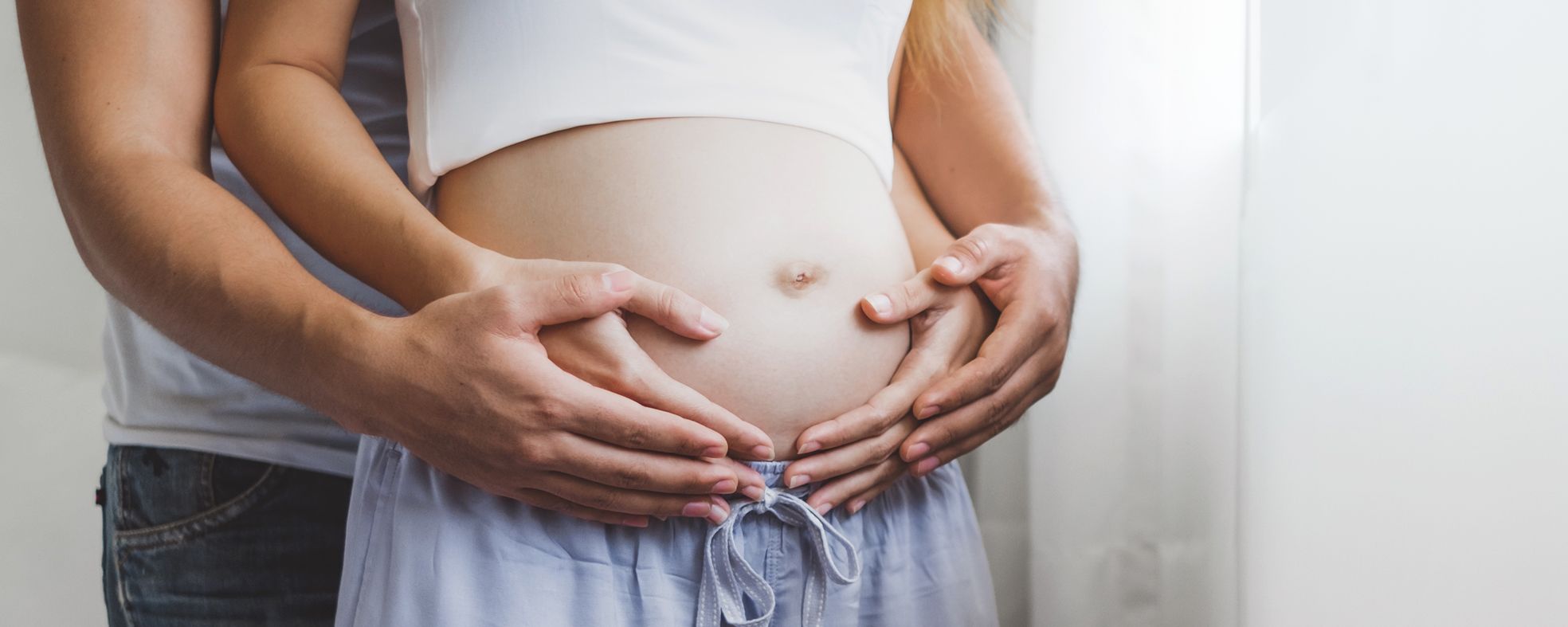 Ein Paar umarmt liebevoll den Bauch einer schwangeren Frau. Ihre Hände sind sanft auf ihrem Bauch platziert, was die Vorfreude auf das kommende Kind symbolisiert. Der Hintergrund ist hell und freundlich, was eine warme Stimmung vermittelt.