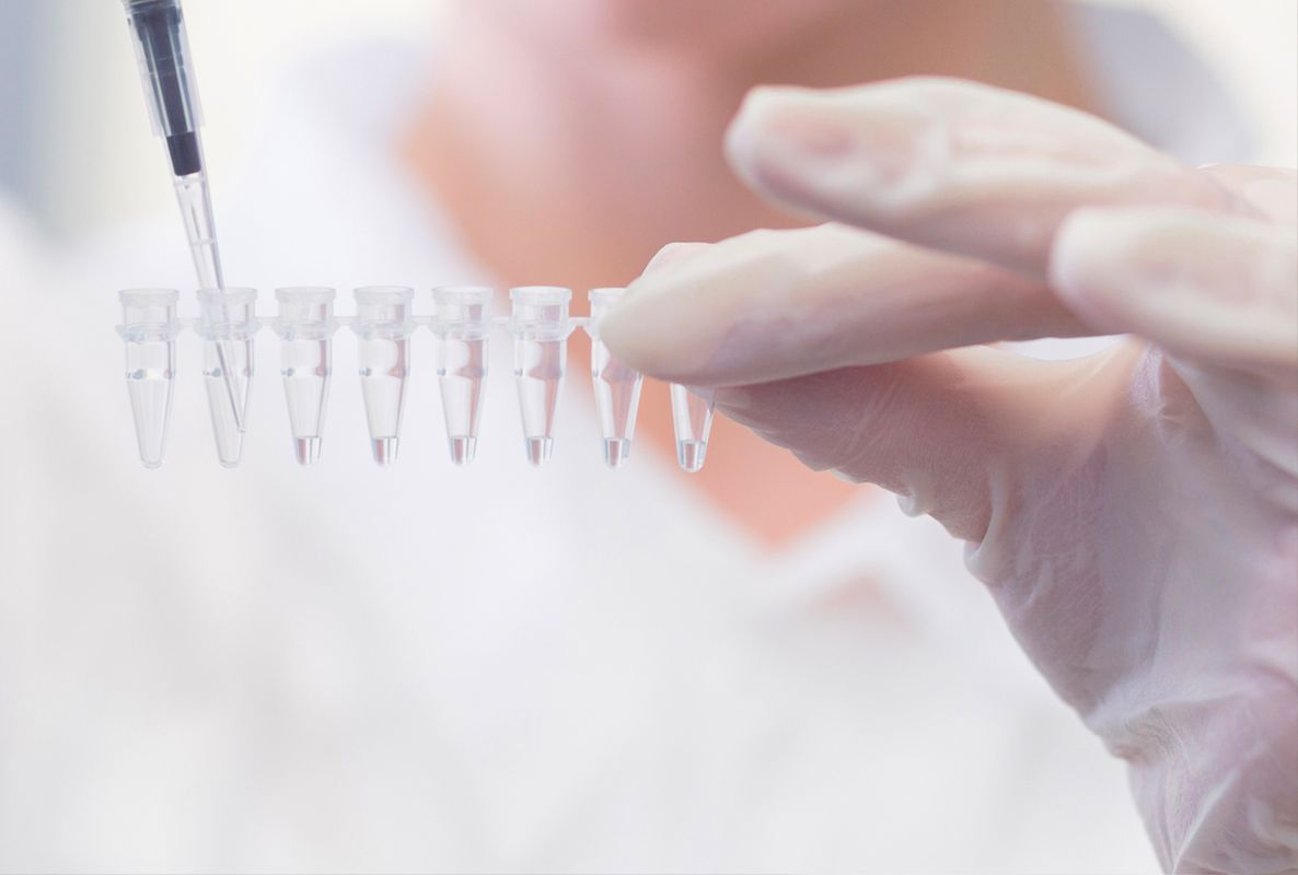 A gloved hand holds a row of clear test tube vials. Another hand is positioned above, using a pipette to transfer liquid into one of the vials. The setting appears to be a laboratory focused on scientific experimentation.