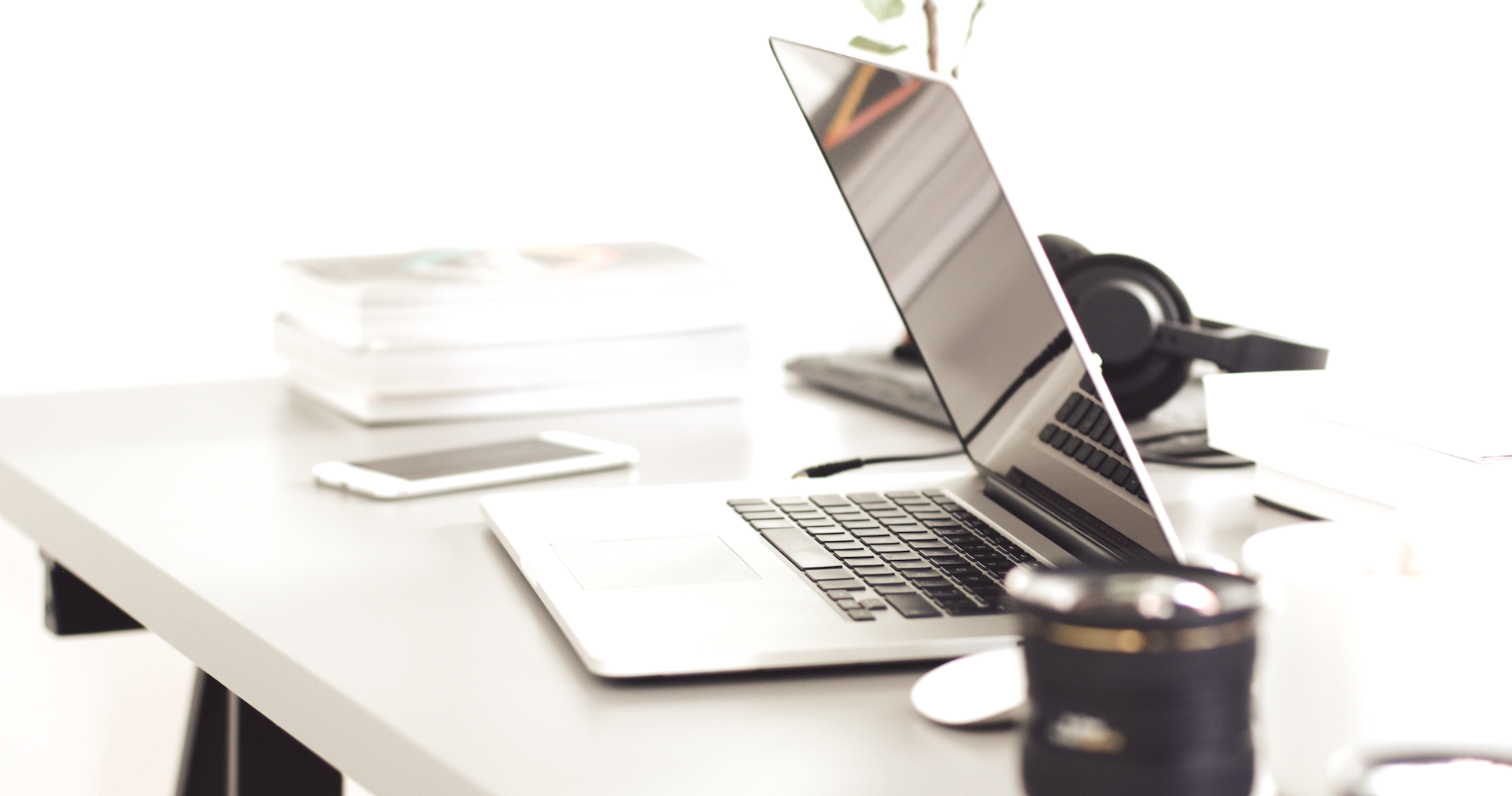 Ein moderner Arbeitsplatz mit einem offenen Laptop, einem Smartphone und einem Becher auf einem Tisch. Im Hintergrund sind Stapel von Papier und Kopfhörer zu sehen, alles in einem hellen, minimalistischen Raum.