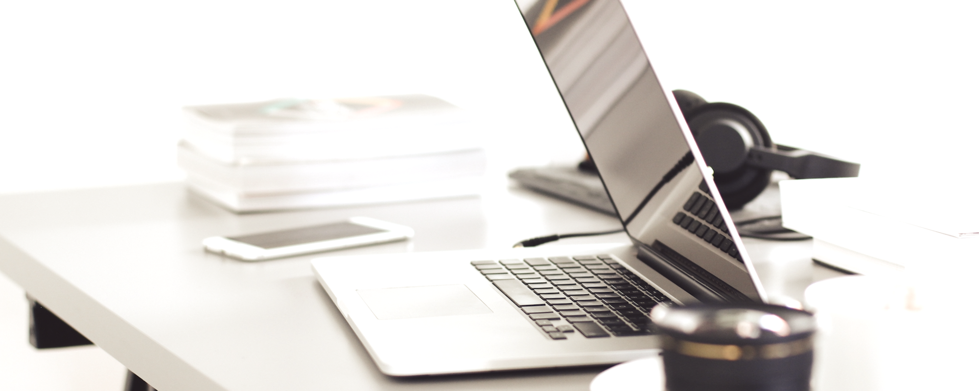 Ein moderner Arbeitsplatz mit einem offenen Laptop, einem Smartphone und einem Becher auf einem Tisch. Im Hintergrund sind Stapel von Papier und Kopfhörer zu sehen, alles in einem hellen, minimalistischen Raum.