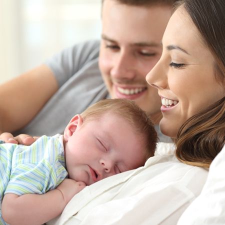 A happy couple smiles as they gently cradle their sleeping baby on a cozy couch. The baby, dressed in a striped outfit, nestles peacefully against one parent's chest, conveying a sense of warmth and tranquility in a loving family moment.
