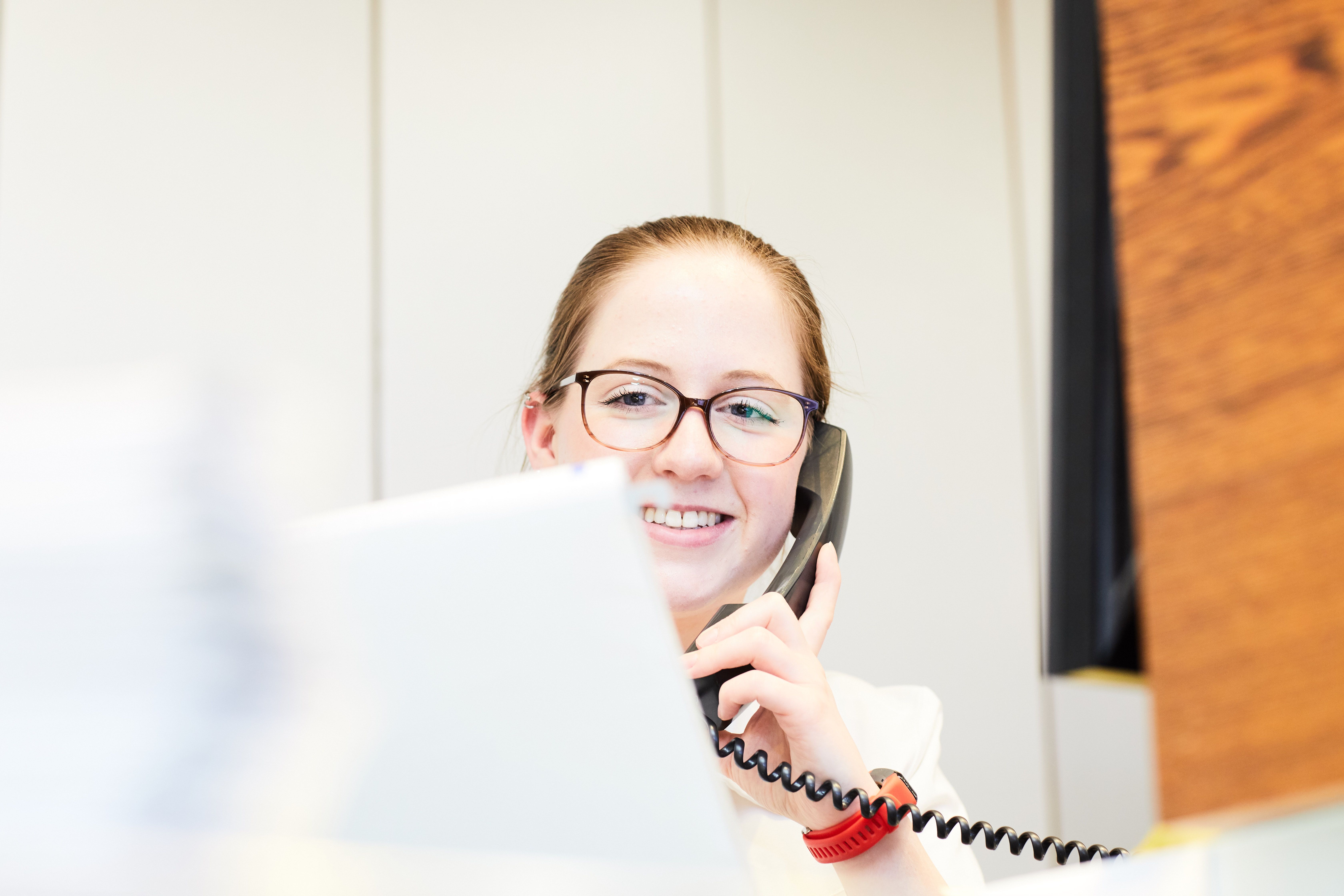 A smiling woman with glasses is on the phone, engaged in conversation. She is seated at a desk with a laptop visible in front of her, suggesting a work environment. The background features light-colored walls and wooden elements.