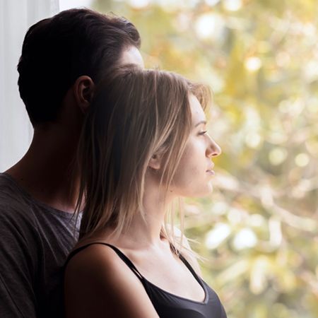 A couple stands by a window, with the woman in the foreground and the man behind her. They both gaze thoughtfully outside, surrounded by soft, blurred greenery, conveying a sense of contemplation and intimacy.