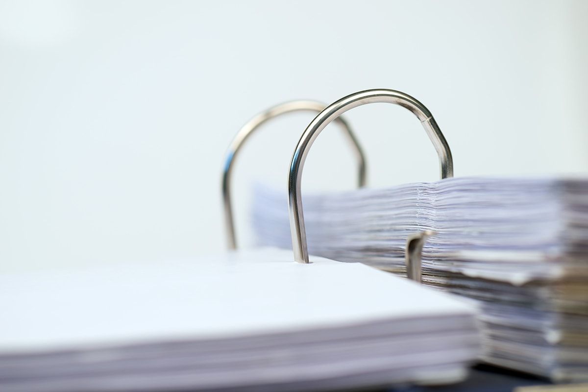 A close-up view of a binder holding a stack of white paper. The binder rings are partially open, highlighting the organized sheets behind them, suggesting an emphasis on documentation or record-keeping. The background appears blurred, focusing attention on the binder and papers.