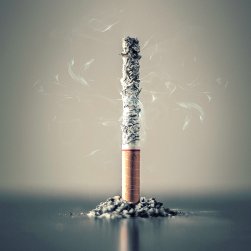 A cigarette butt, predominantly ash and burnt paper, sits upright among a pile of ash, with wisps of smoke rising from the top. The background is a soft gradient, emphasizing the cigarette's solitary presence.