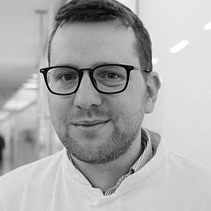 Ein junger Mann mit Brille und einem weißen Kittel lächelt in die Kamera. Er steht in einem hellen, modernen Innenraum mit verschwommenen Personen im Hintergrund. Seine Frisur ist kurz und leicht unordentlich.