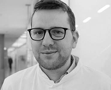 A man with short hair and glasses smiles at the camera, wearing a white coat. The background features a blurred interior, suggesting a clinical or professional environment.