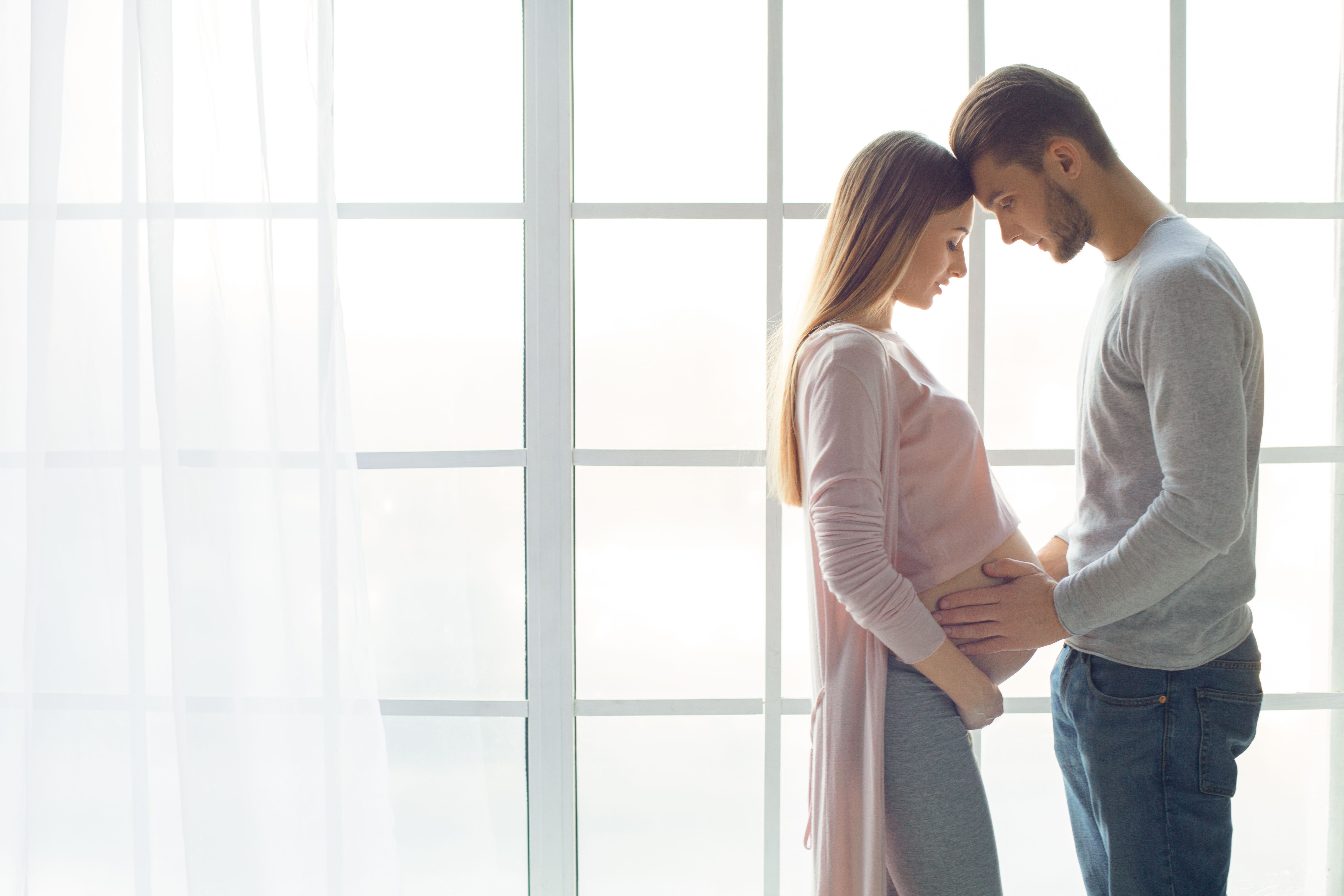 A couple stands together in a softly lit room, the woman is pregnant and gently cradling her belly while the man leans in, smiling at her. They share a tender moment, reflecting love and anticipation for their upcoming child.