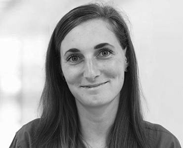 A smiling young woman with long, dark hair and light skin is facing the camera. She has a subtle expression and is wearing a simple top. The background is softly blurred, emphasizing her face.