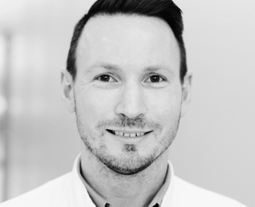 A close-up, black and white portrait of a smiling man with short hair and a trimmed beard. He is wearing a light-colored collared shirt and is looking directly at the camera, conveying a friendly demeanor.