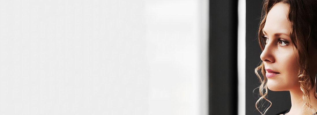 A close-up profile of a woman with wavy hair, looking thoughtfully out of a window. The background is softly blurred, focusing attention on her contemplative expression. The lighting highlights her features and creates a serene atmosphere.