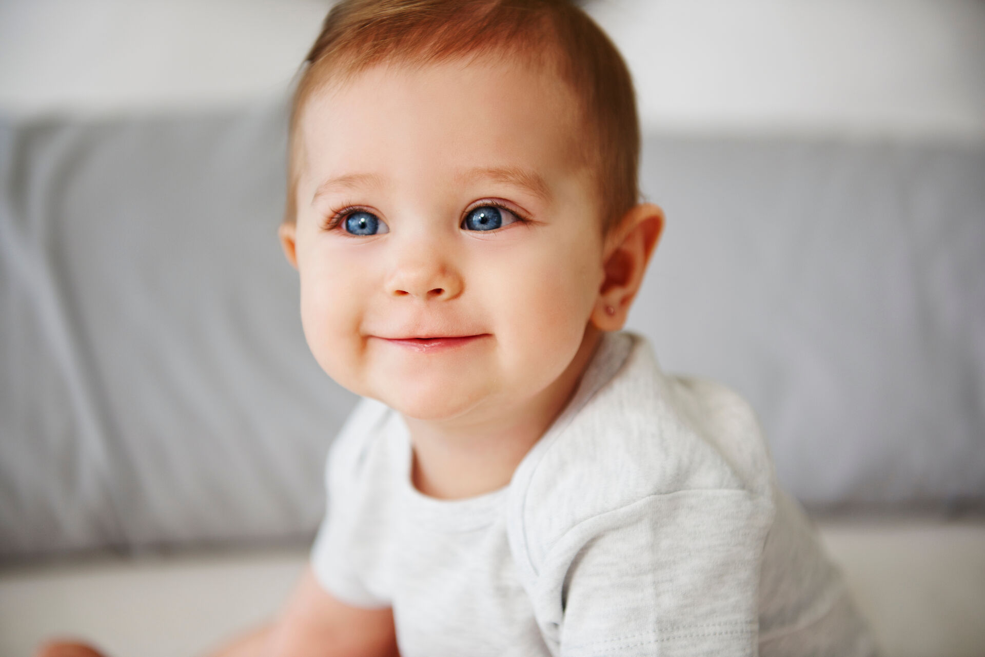 Ein lächelndes Baby mit blauen Augen trägt ein graues Oberteil. Es sitzt auf einem Bett und schaut in die Kamera, dabei strahlt es Freude aus. Die Umgebung wirkt weich und einladend.