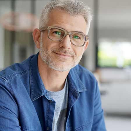 A middle-aged man with short, gray hair and a beard smiles warmly while wearing glasses and a denim jacket. He appears relaxed and approachable, with a soft background that suggests a modern indoor setting.