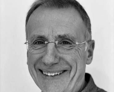 A smiling older man with short hair is wearing glasses. He has a friendly expression, showcasing a warm demeanor. The image is in black and white, focusing on his face and upper shoulders against a plain background.