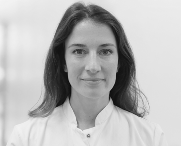 A woman with long, dark hair is wearing a white medical coat. She has a neutral expression and is facing the camera directly. The background is blurred, emphasizing her. The overall tone of the image is professional and clinical.